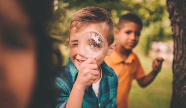 
		Portrait of a little boy holding a large round magnifying glass to his face, making his eye look humorously large as he plays with friends in a summer park.
	