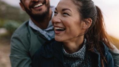 
		Young couple outdoors laughing together. He holds her from behind.
	