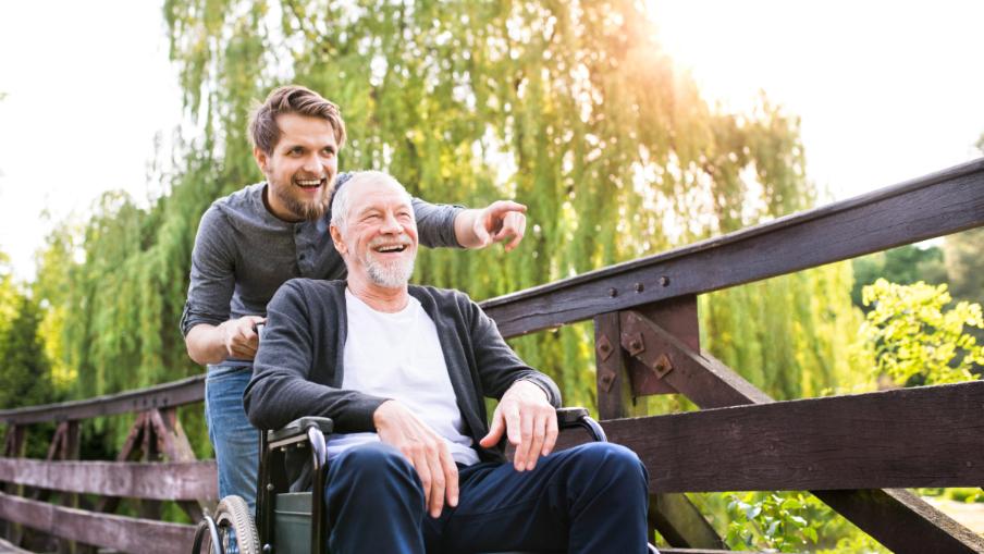 
		Young man standing on a bridge with an older man in a wheelchair, both laughing.
	