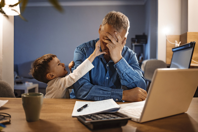 A stressed father sits at his laptop with his son