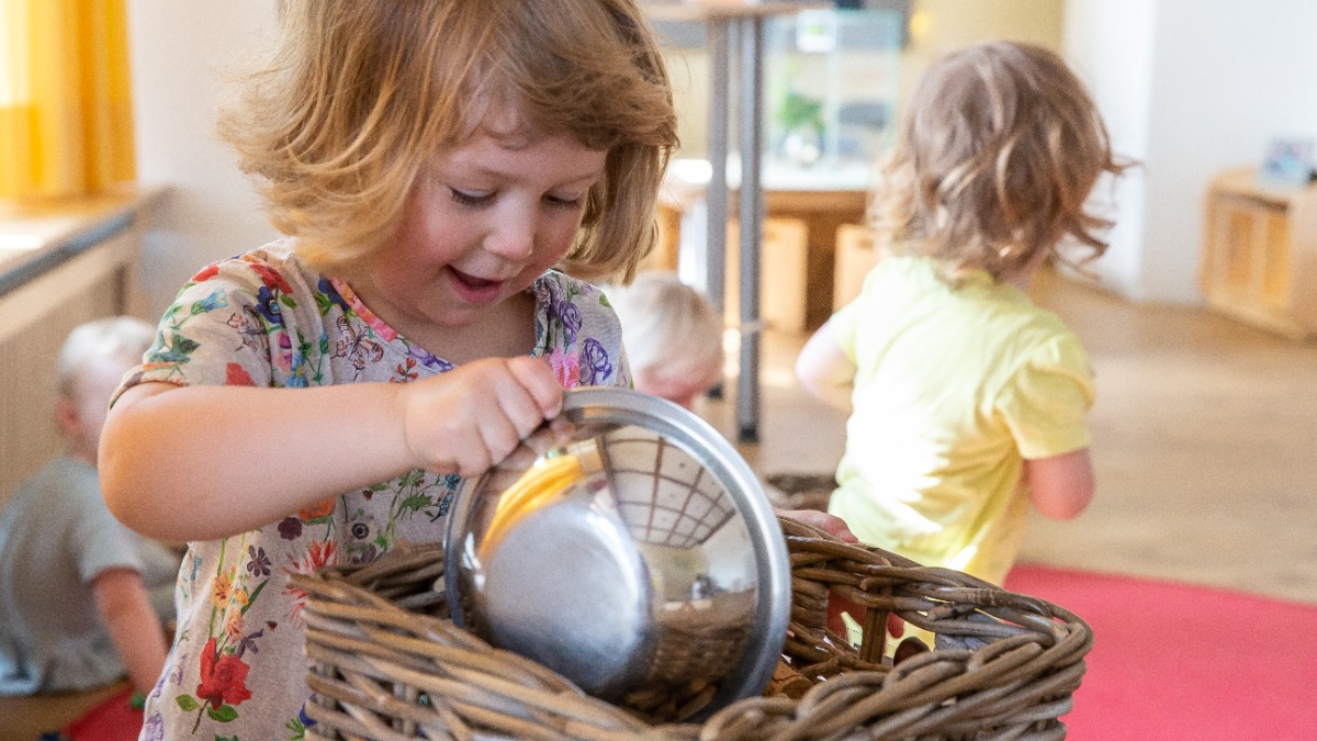 Approximately three-year-old girl at childcare center holds a basket and a metal bowl in her hands