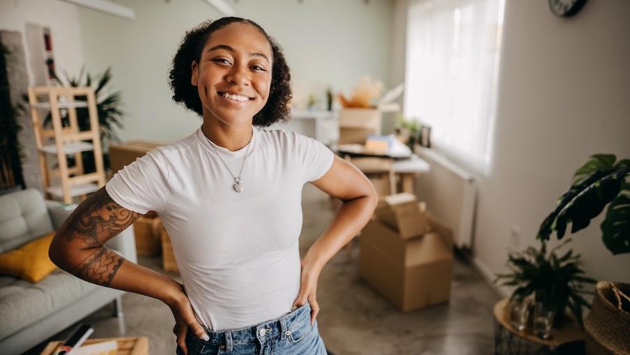 
		A young woman stands in her first apartment of her own, with boxes behind her
	