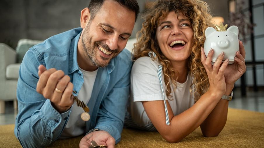 
		A man and a woman are sitting at the table. She is happily shaking the piggy bank and he has coins in his hand and is laughing.
	