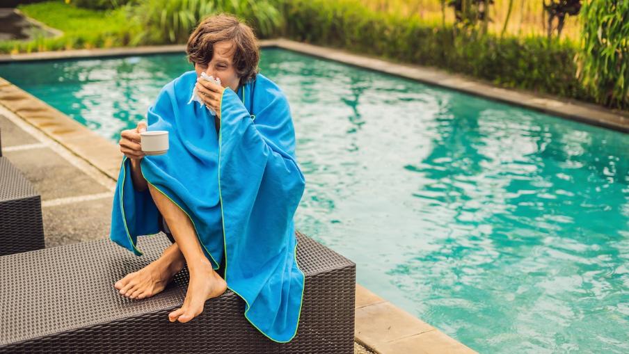 
		A young man sits by the pool with a blanket wrapped around him and blows his nose
	