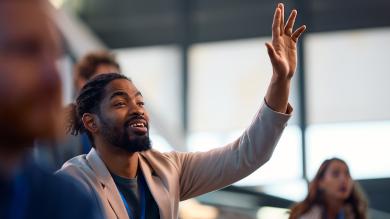 
		A young man stretches his hand up and answers. He is sitting among other colleagues in the office.
	