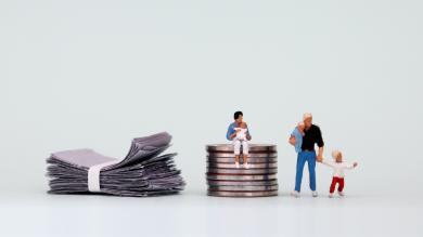 
		A miniature woman with a baby sits on a pile of coins next to a pile of bills and a miniature man with a baby next to them.
	