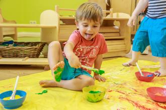 
		A child is sitting on the floor at childcare center . There are paint buckets and brushes around him and the child is painting his foot with blue paint.
	