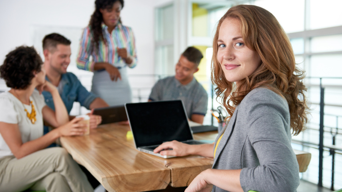 EIne Vereinbarkeitsmanager sitzt an einem Tisch mit ihrem Laptop und lacht in die Kamera. Ihre Kollegen sind verschwommen im Hintergrund zu sehen.