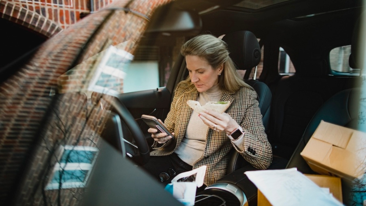 A woman eats a sandwich in the car while typing on her smartphone.