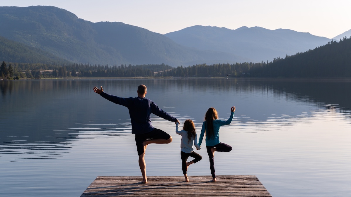 Father stands with his two children on one leg on a jetty by the lake