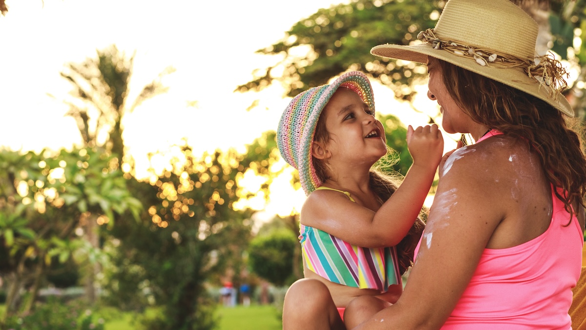 Happy mother sitting in the sunshine under a tree with her toddler and applying sunscreen