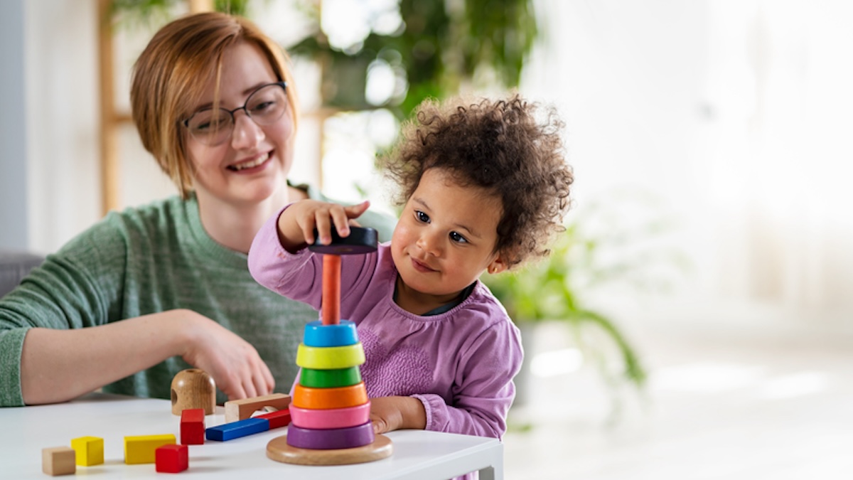 A small child plays with building blocks at a small table and his mother sits smiling next to him