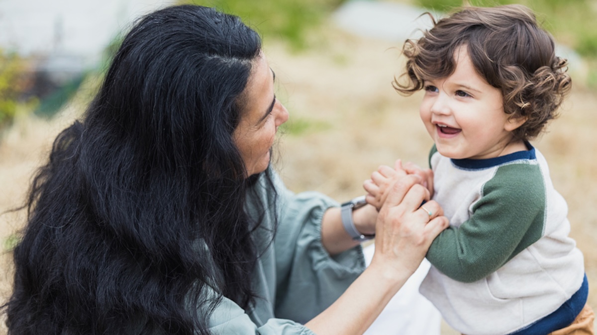 Mother crouches in front of her small child and grabs his arms, child looks happily to the side