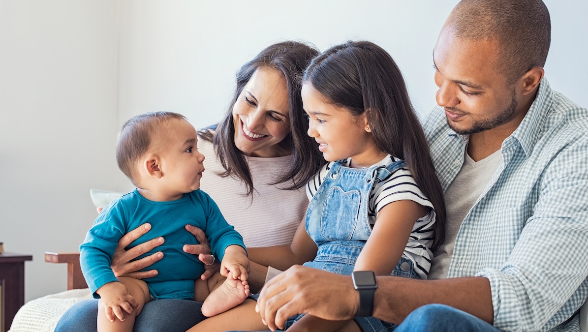 Parents sit on the sofa laughing with their two toddlers