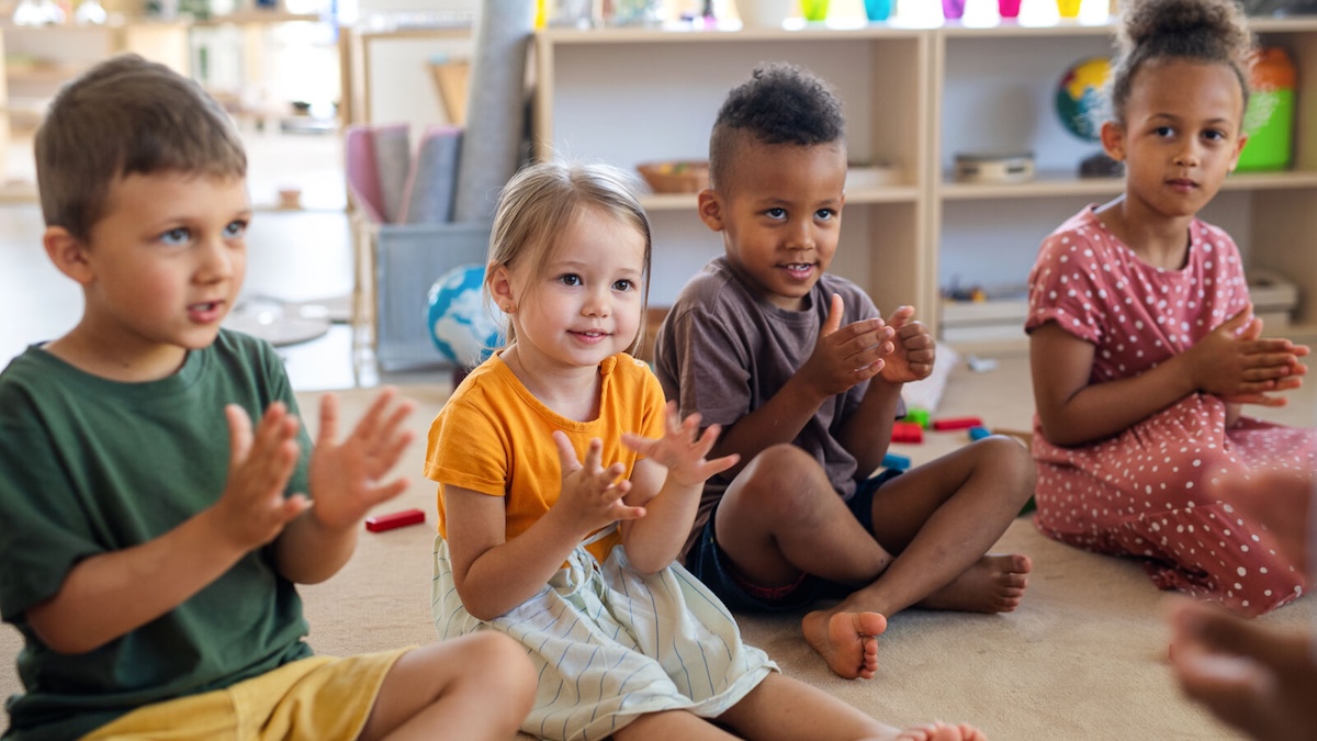 Four toddlers are sitting next to each other, clapping and looking at their caregiver