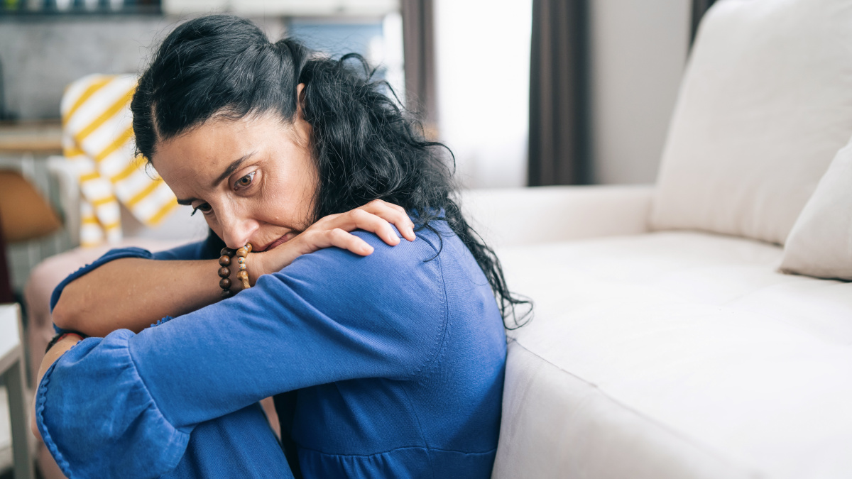 A woman sits on the floor at home and looks down anxiously