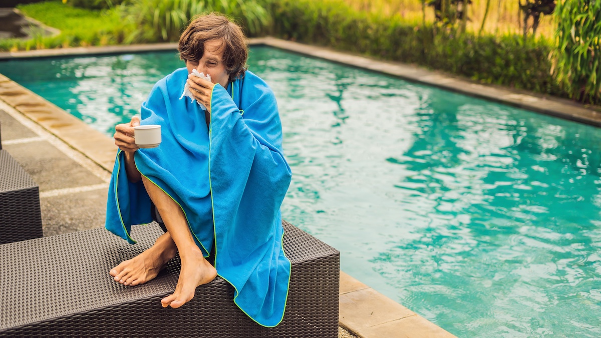 A young man sits by the pool with a blanket wrapped around him and blows his nose